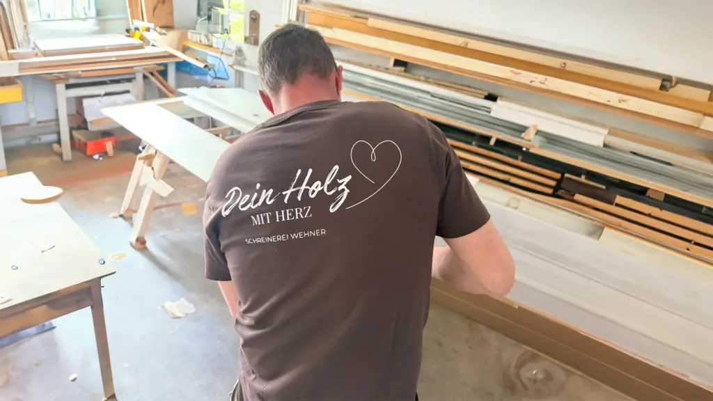 Schreiner Sebastian Wehner bei der Arbeit. Er trägt ein T-Shirt mit dem Firmennamen "Dein Holz mit Herz" auf dem Rücken